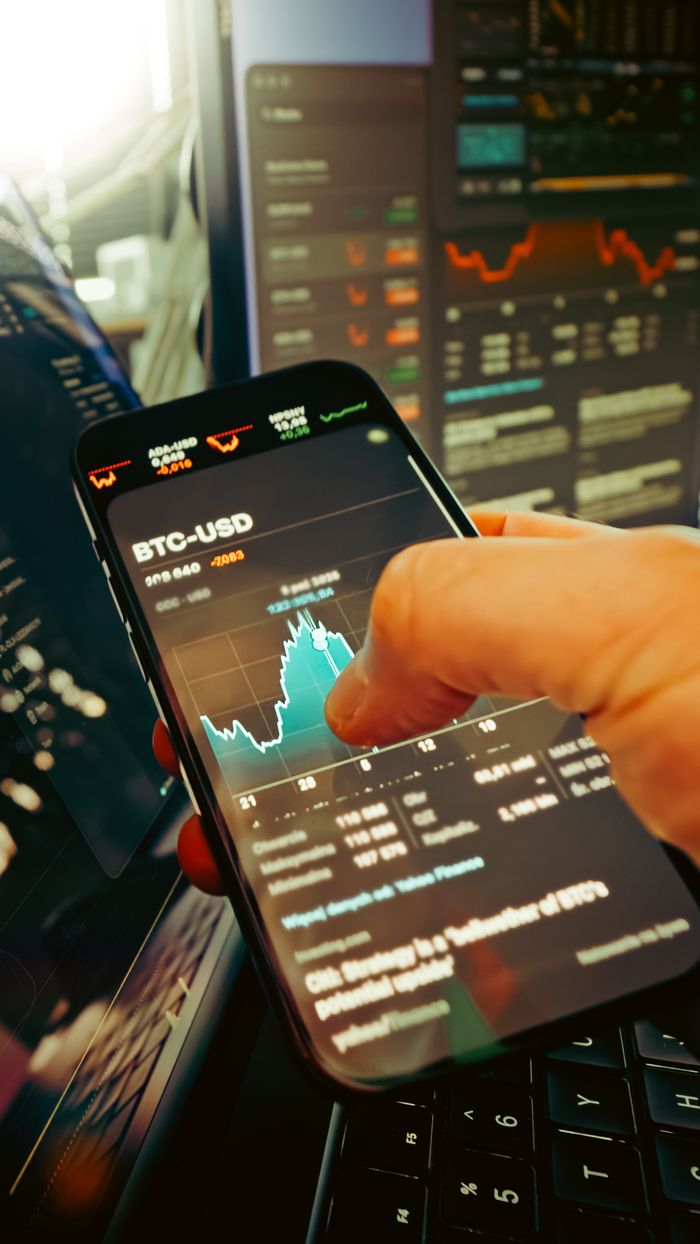 creative-01 Une main tient un smartphone affichant le graphique BTC-USD au milieu d’écrans de trading numériques, mettant en avant la finance crypto.